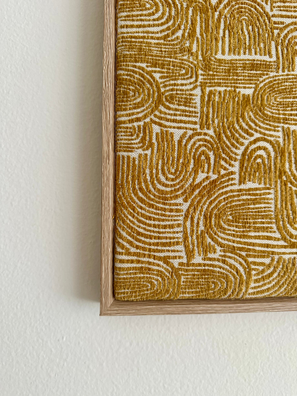 Close-up of a framed wall art piece showing an abstract wavy line pattern in mustard gold and off-white, mounted on a light neutral wall.