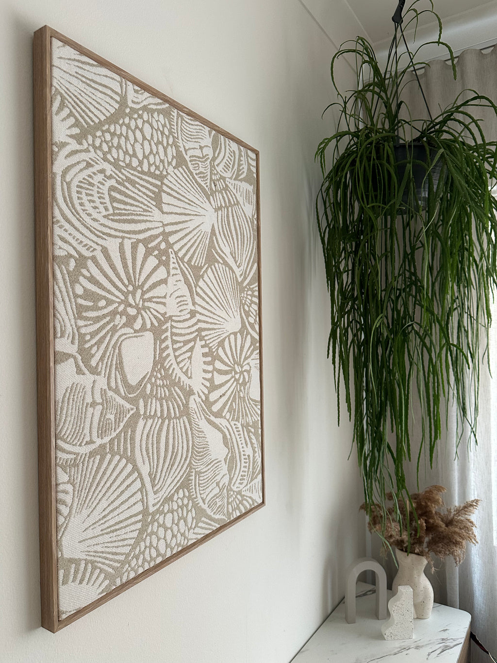 A rectangular framed art print featuring a detailed line art pattern of seashells and starfish in beige on a white background, styled on a white surface with a green plant in the background.