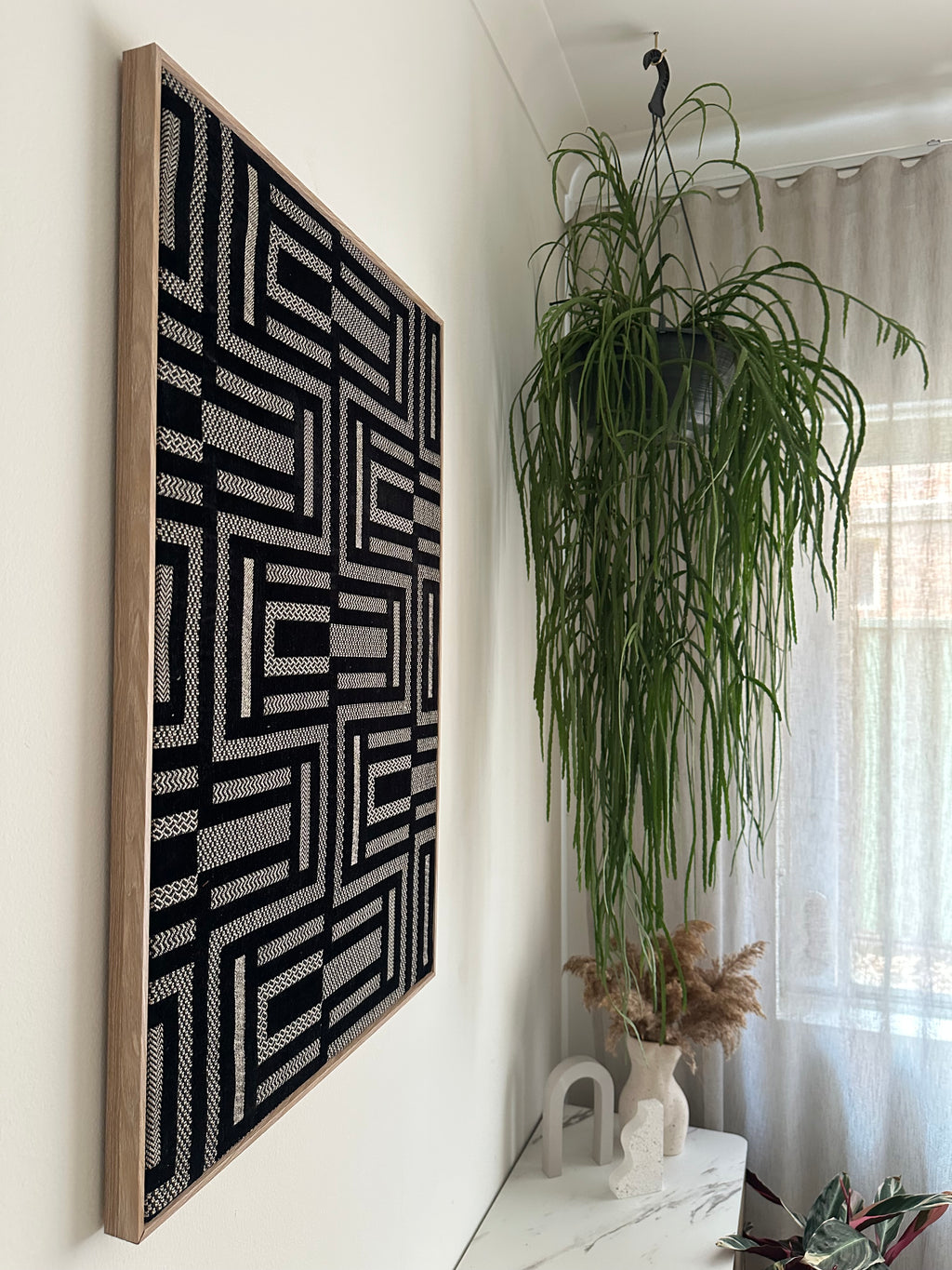 A large framed wall art piece with a black and white geometric maze-like pattern hangs on a white wall in a bright room next to a tall green hanging plant.