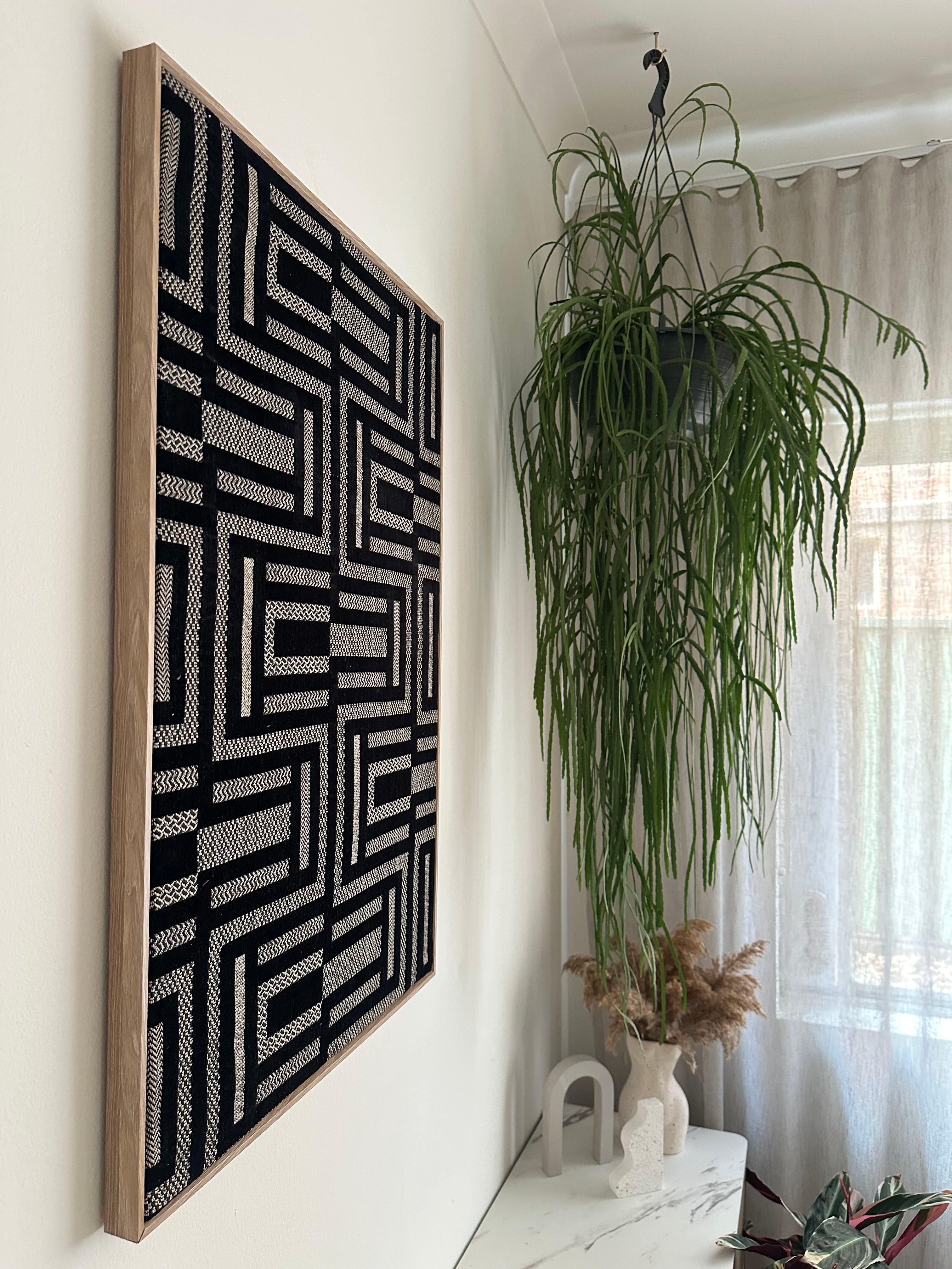 A large framed wall art piece with a black and white geometric maze-like pattern hangs on a white wall in a bright room next to a tall green hanging plant.