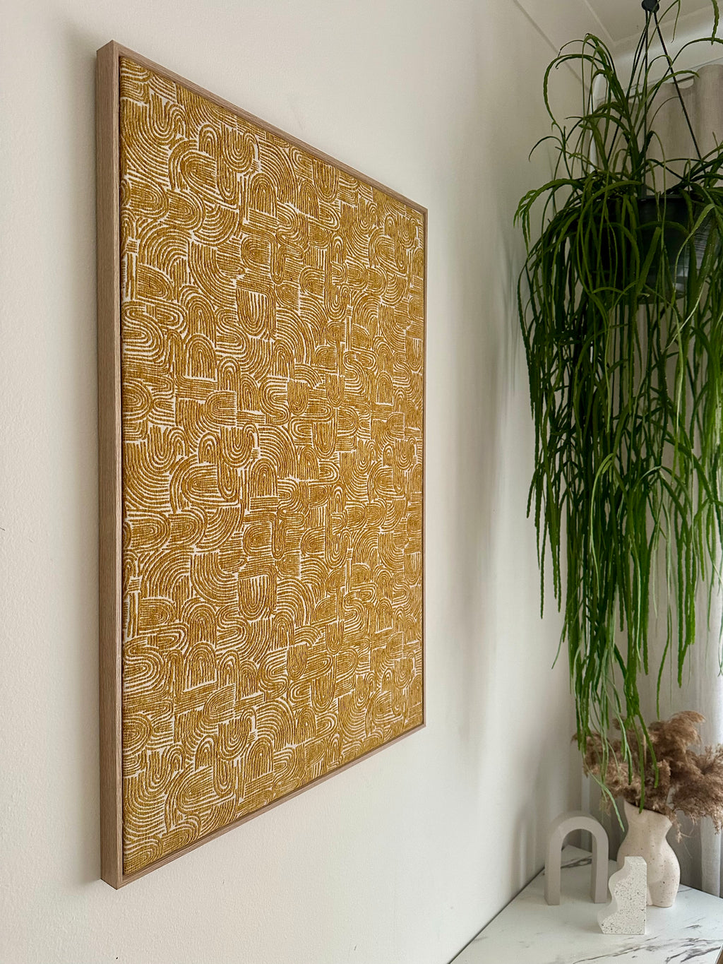 A large framed abstract canvas print with a textured mustard yellow background and white hand-drawn line patterns, hanging on a white wall next to a green potted plant.
