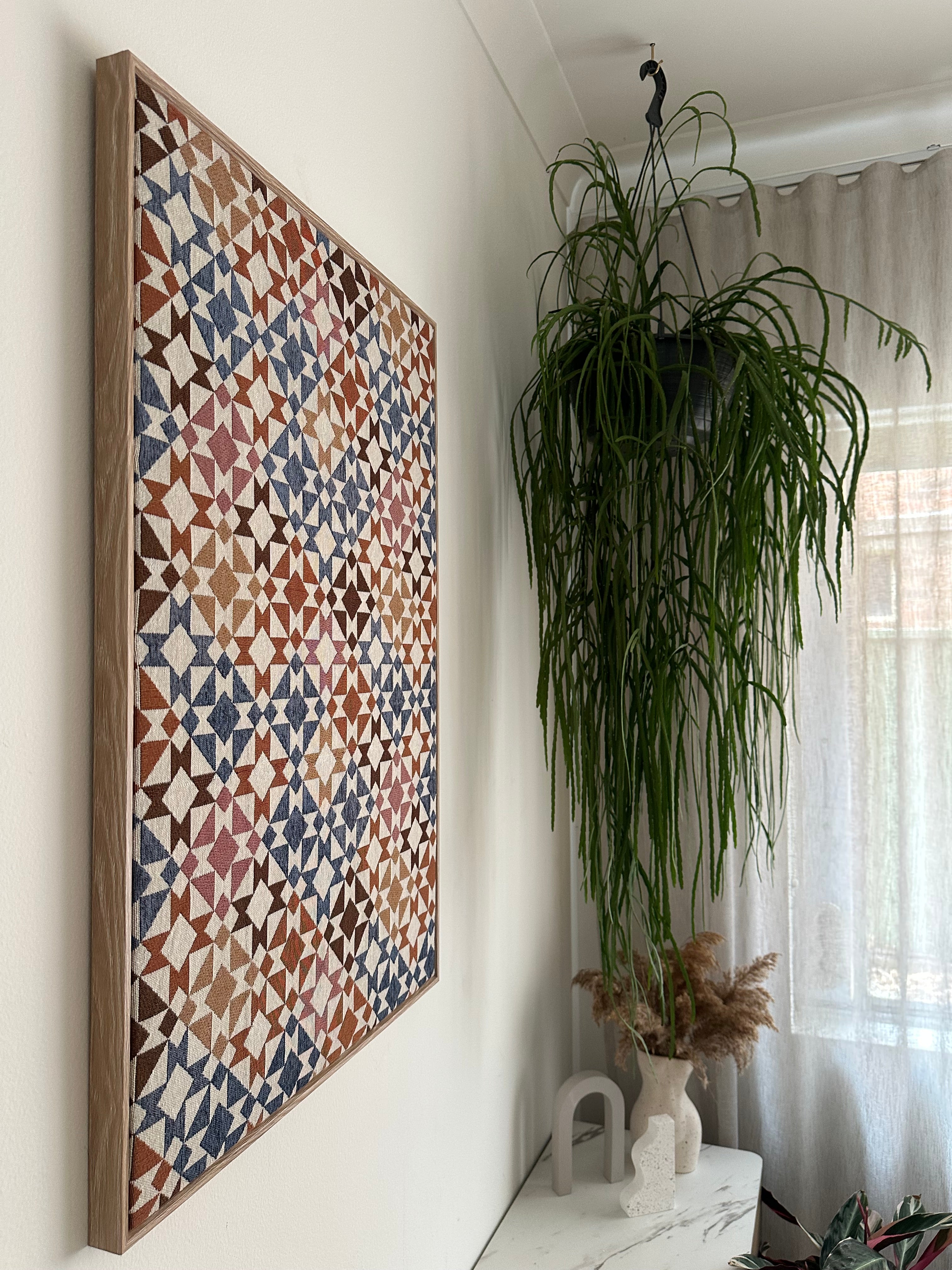 A rectangular wooden tray with a colorful geometric mosaic pattern, displayed on a white surface with a potted green plant and a white ceramic object in the background.