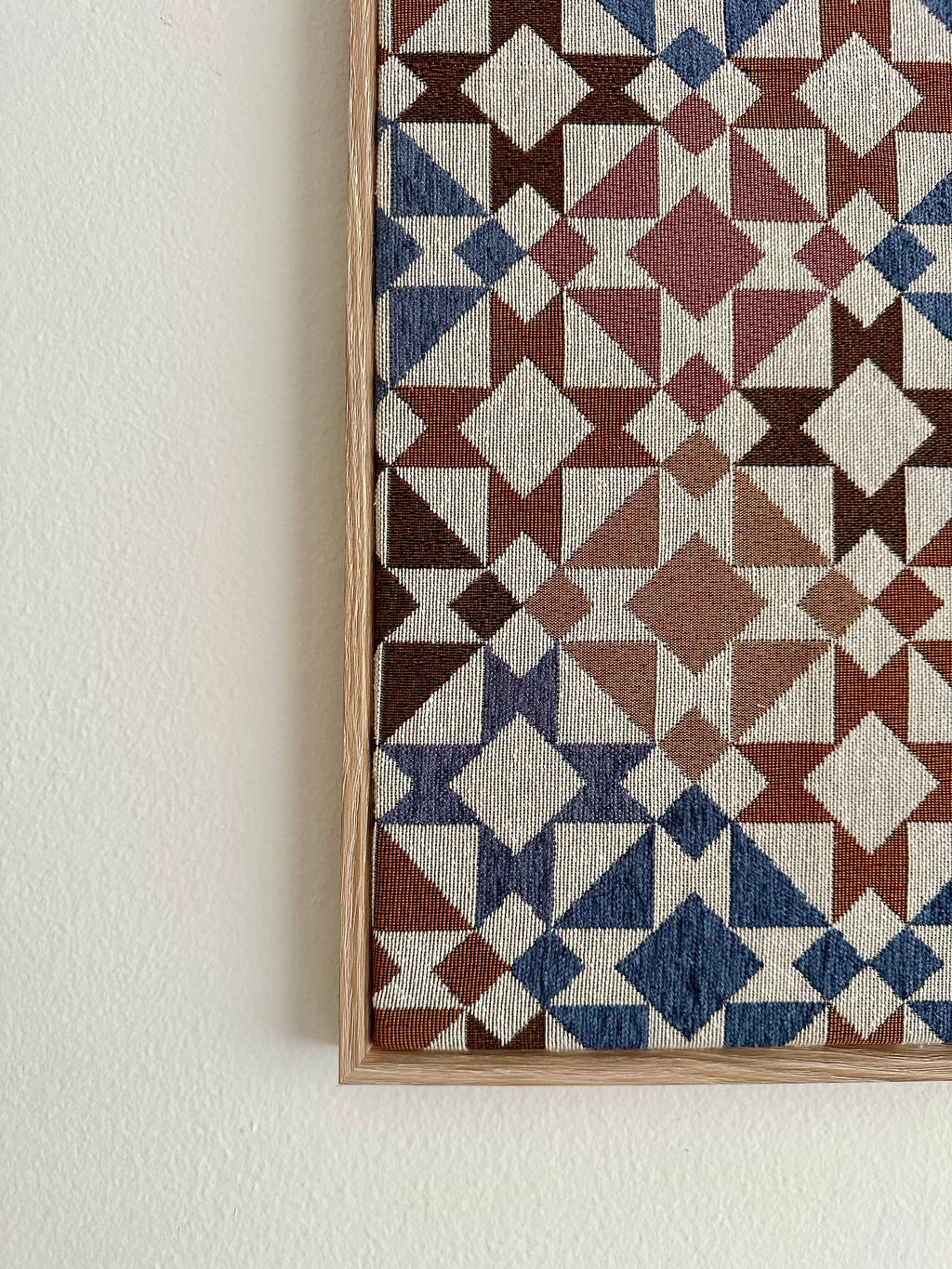 A close-up of a framed textile wall hanging with a geometric pattern in cream, blue, rust brown, and dark brown, mounted on a white wall.