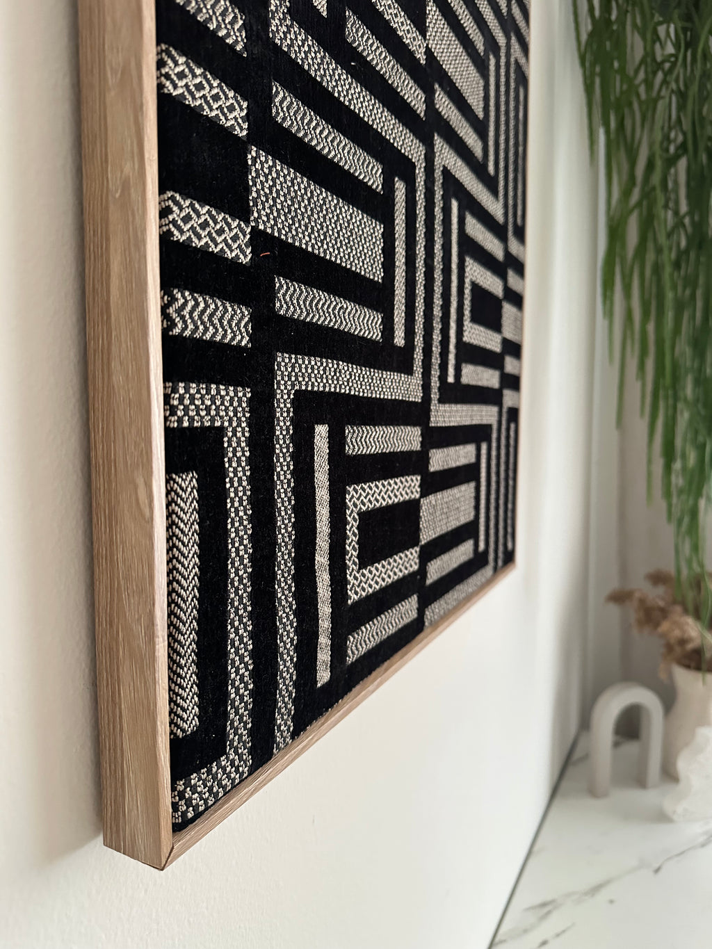 A square woven textile piece with a black and off-white geometric maze-like pattern, finished with a light natural wood frame, resting on a white surface.
