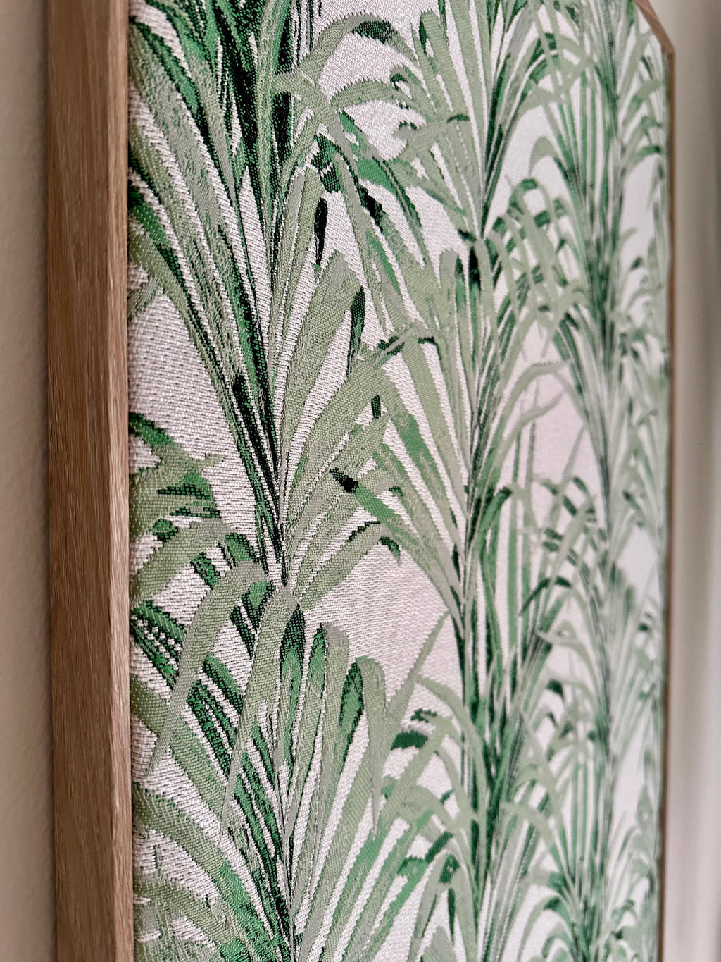 A close-up of framed botanical wall art featuring a green palm leaf pattern on a light background, with a natural wood frame visible on the left.