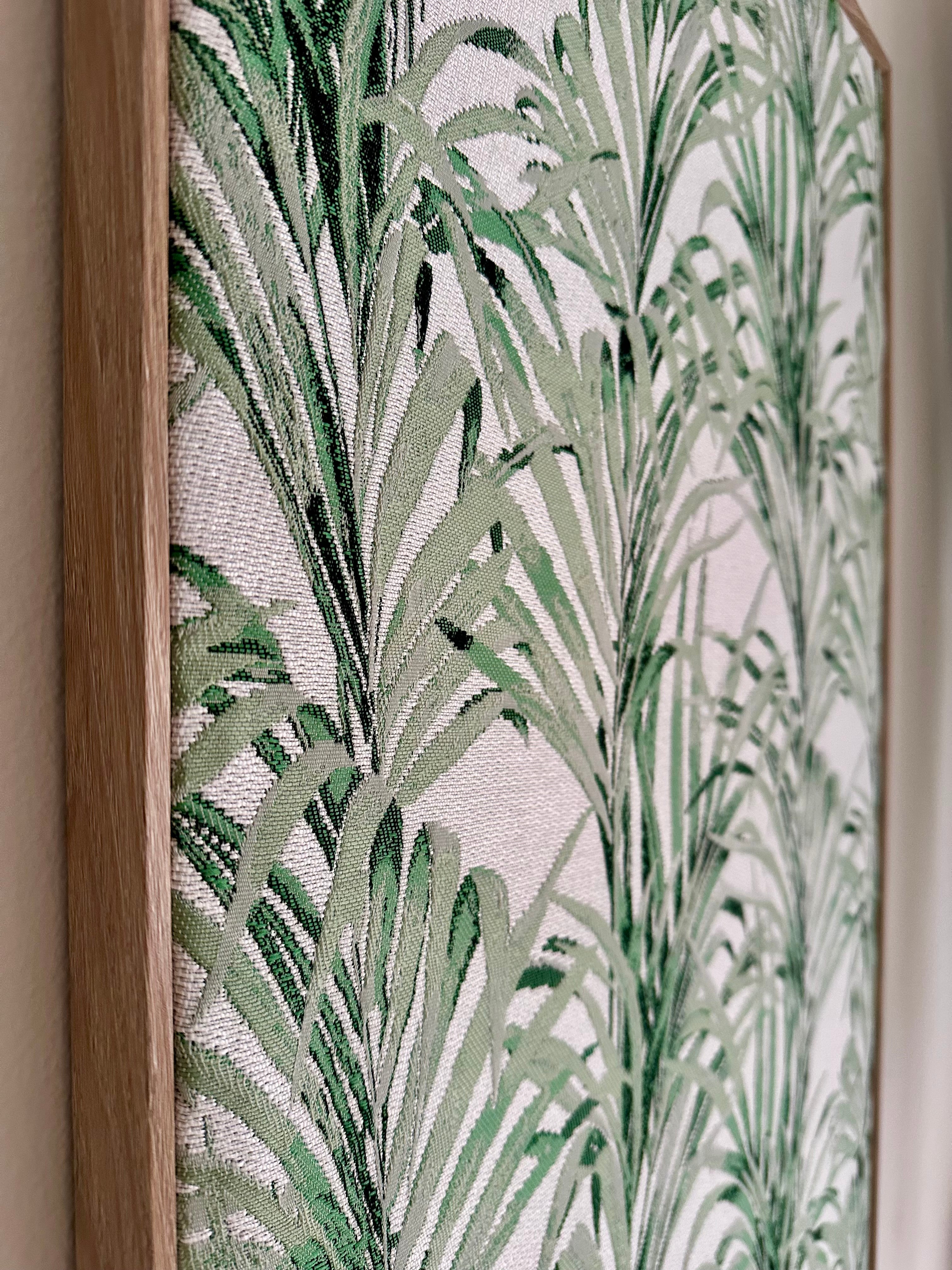 A close-up of framed botanical wall art featuring a green palm leaf pattern on a light background, with a natural wood frame visible on the left.