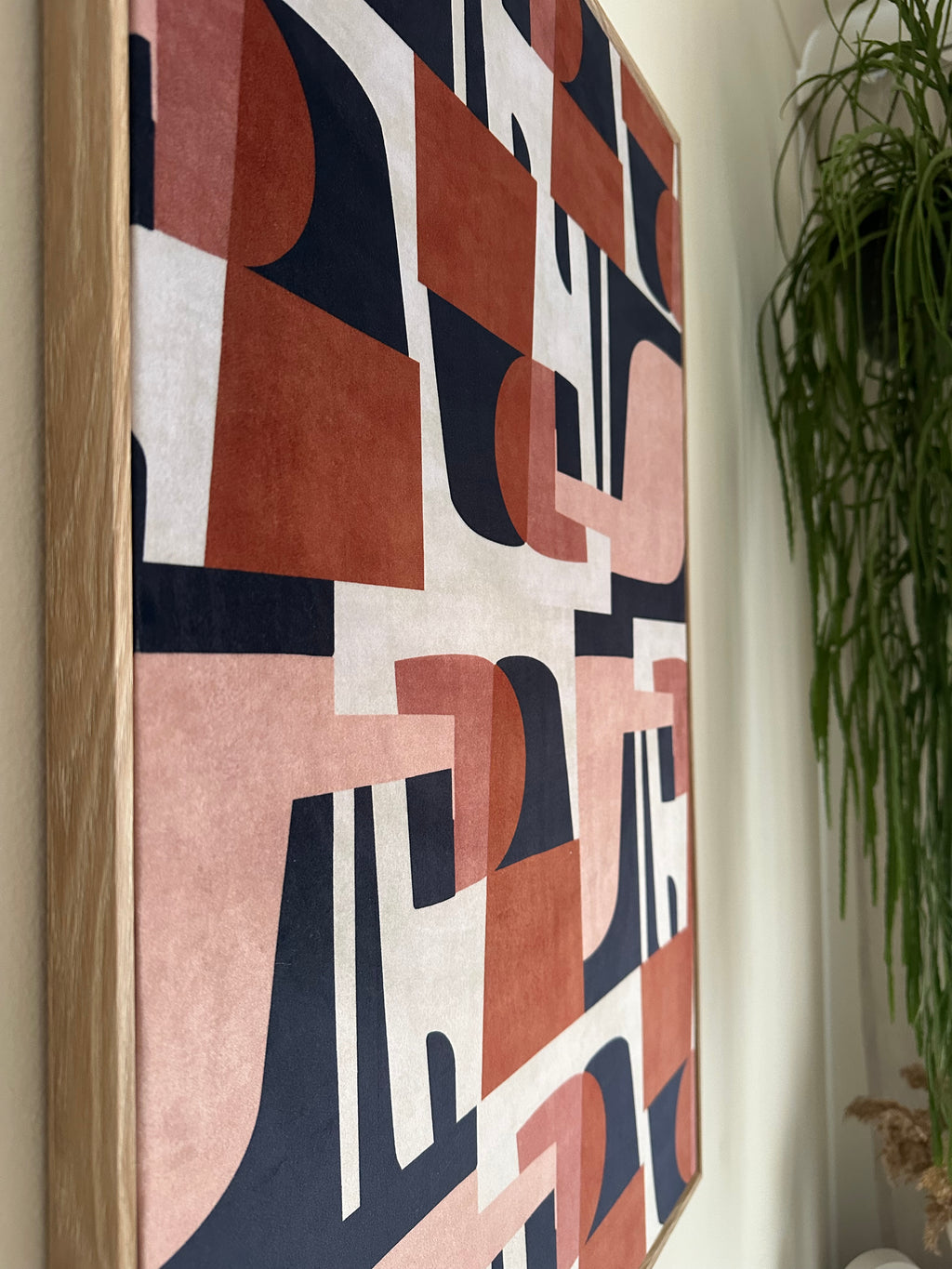 A rectangular framed canvas print featuring a modern abstract geometric pattern in terracotta, dusty pink, navy blue, and off-white, resting on a light surface with green foliage in the background.