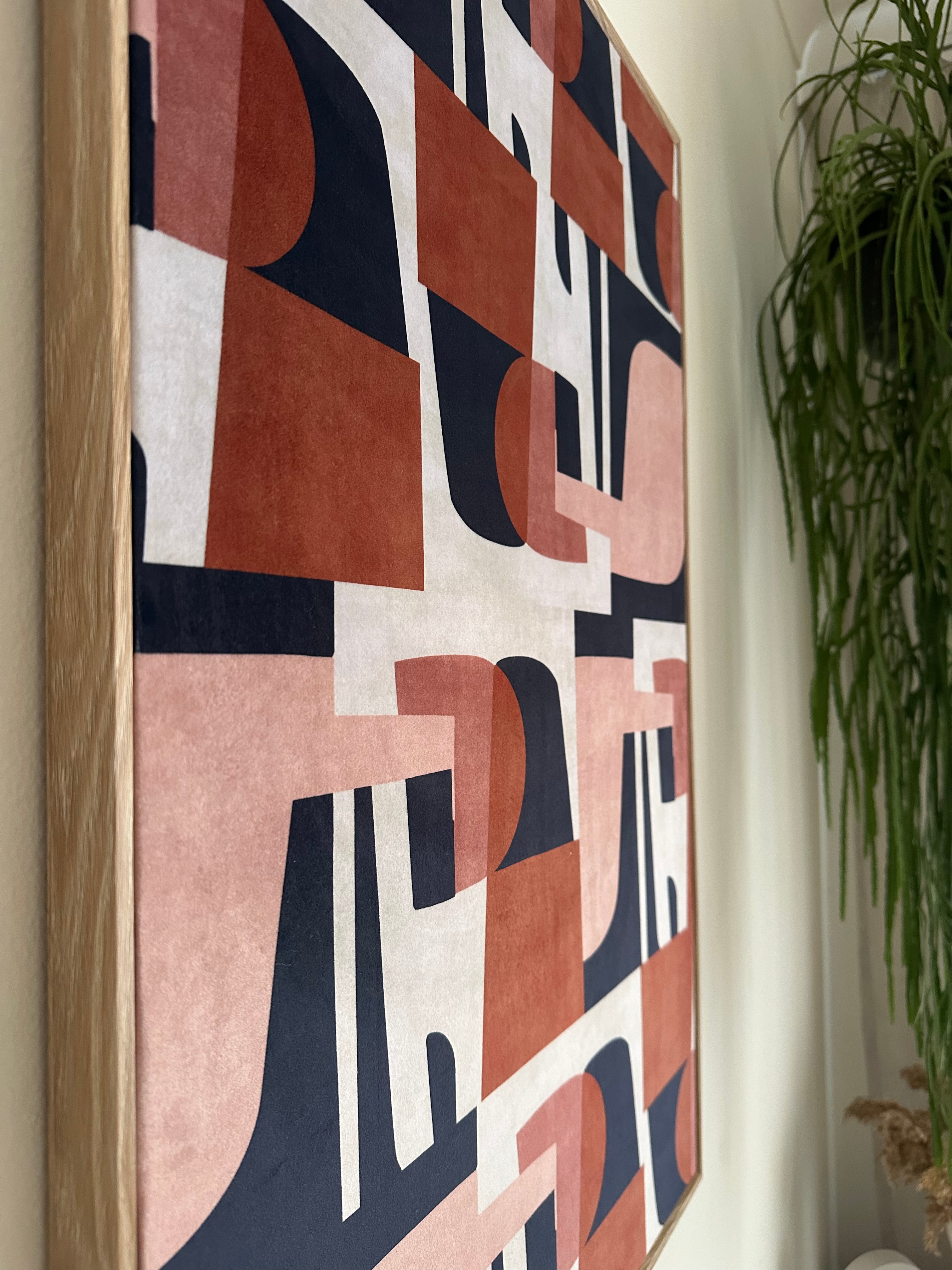 A rectangular framed canvas print featuring a modern abstract geometric pattern in terracotta, dusty pink, navy blue, and off-white, resting on a light surface with green foliage in the background.