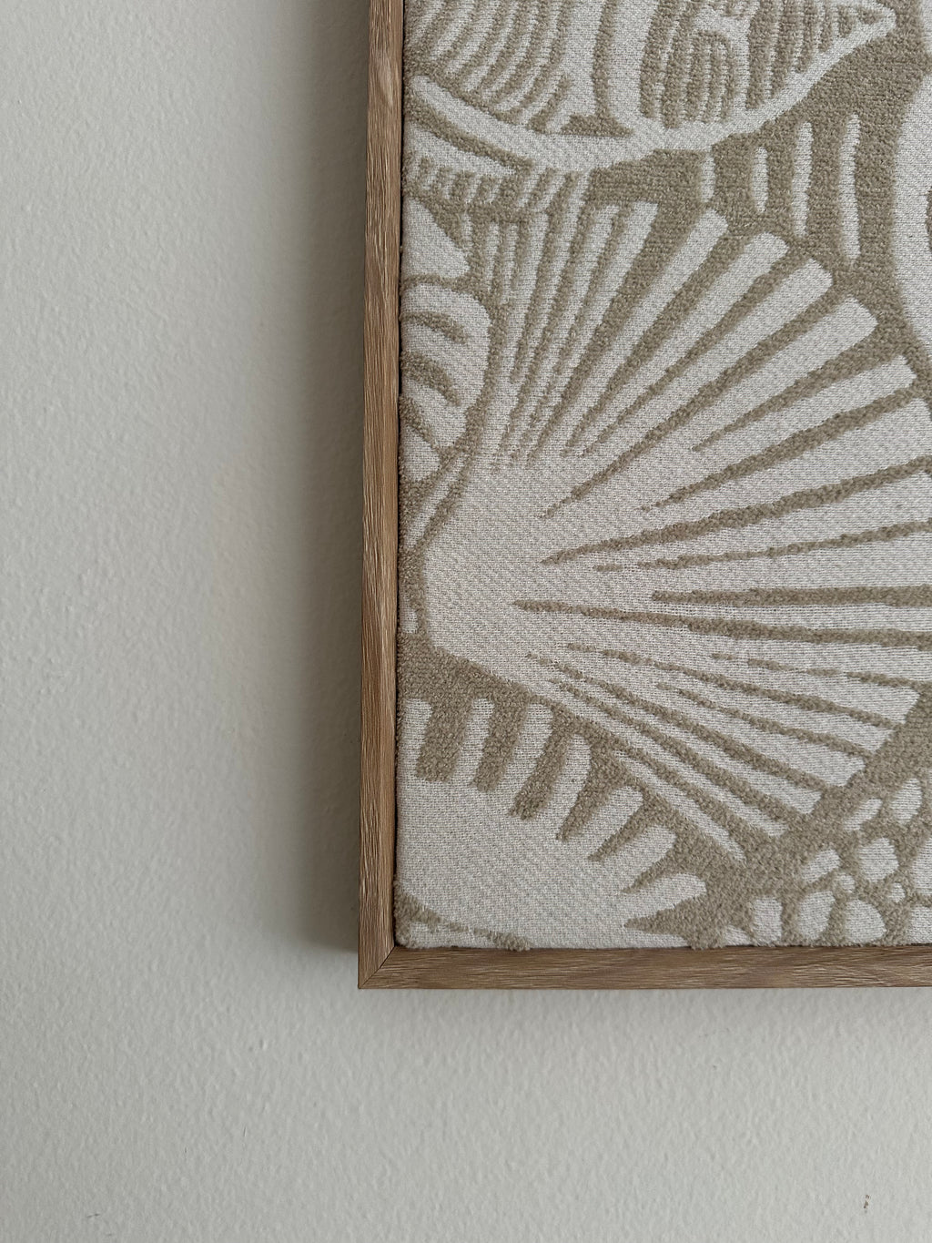 Close-up of a framed textile wall hanging with a woven shell-like pattern in beige and off-white, mounted on a light neutral wall.