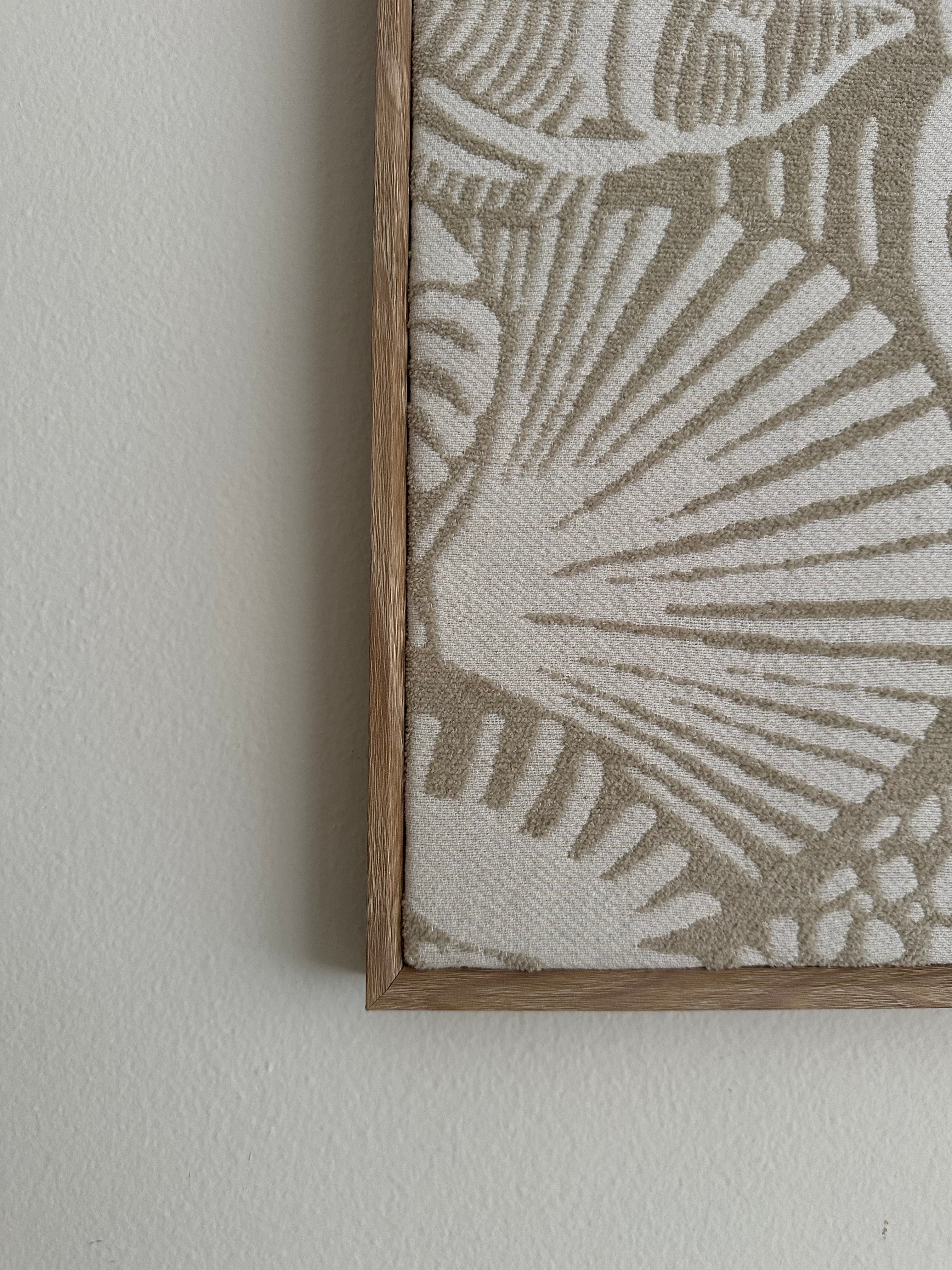 Close-up of a framed textile wall hanging with a woven shell-like pattern in beige and off-white, mounted on a light neutral wall.