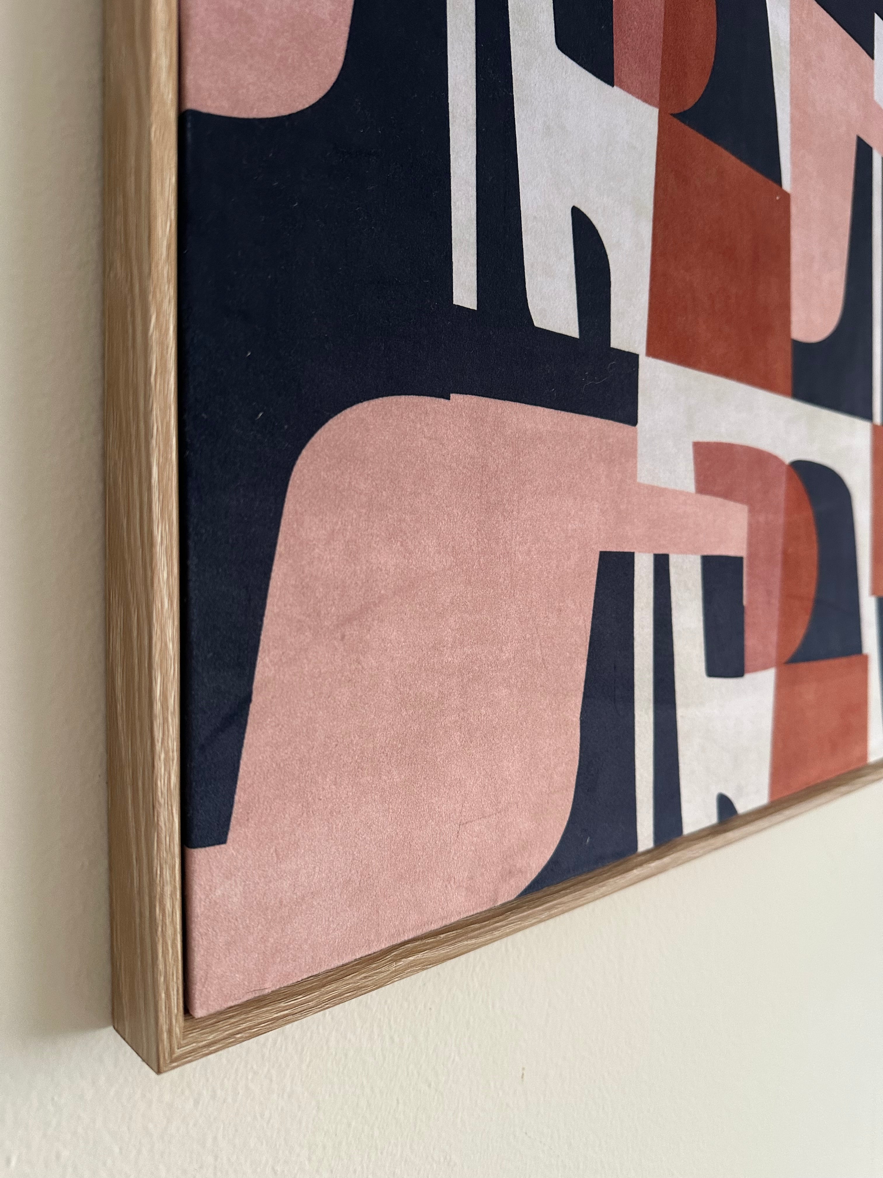 Close-up of a framed abstract wall art print with geometric shapes in blush pink, terracotta, cream, and navy on a light wood frame, mounted on a white wall.