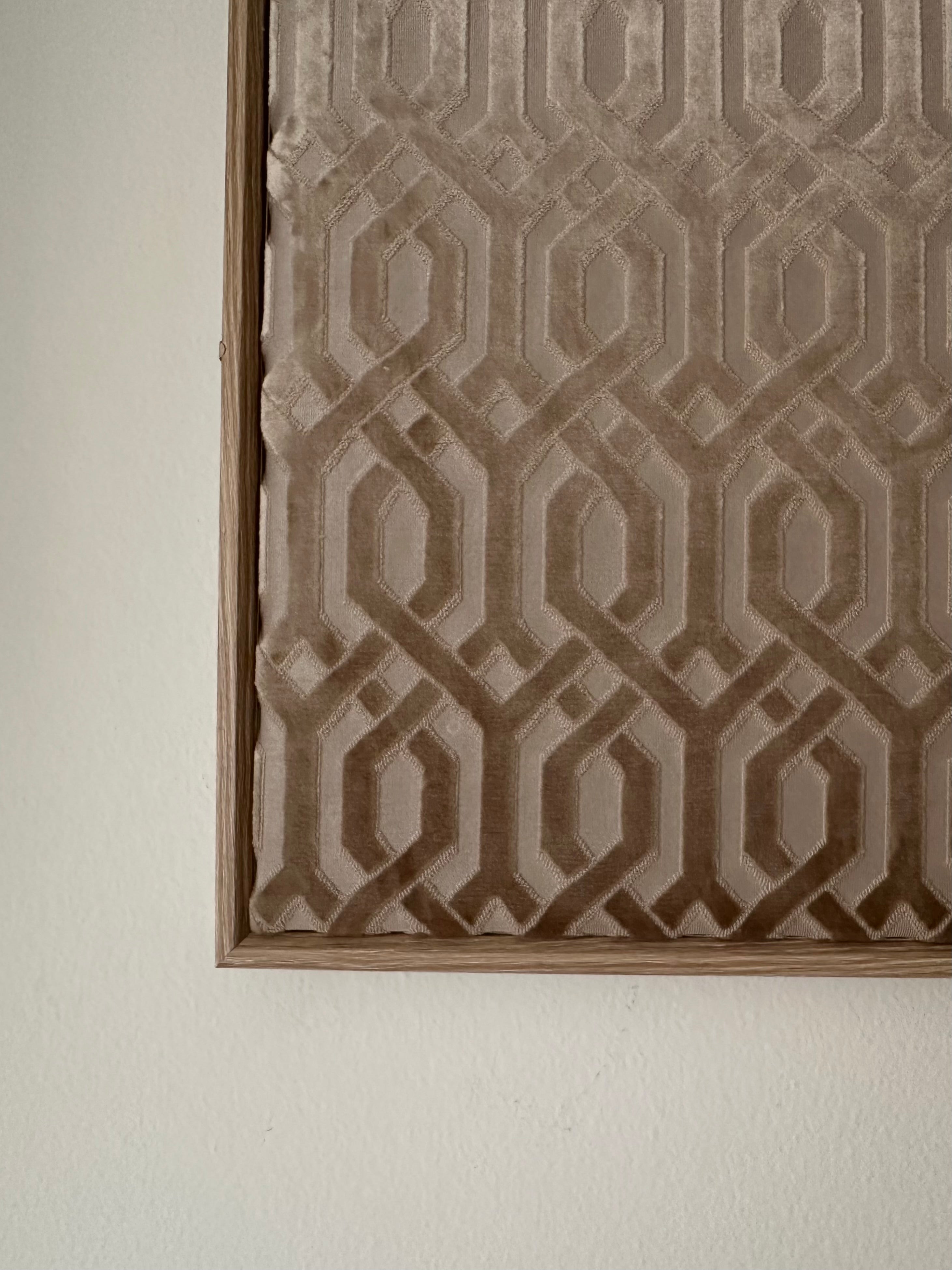 A close-up of a framed wall art piece featuring a geometric interlocking pattern in taupe and brown tones, mounted on a plain off-white wall.