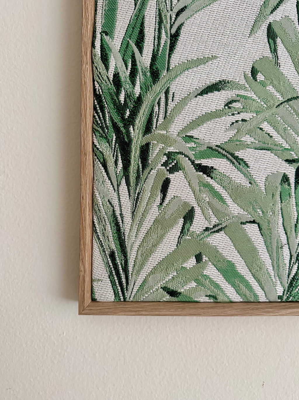A close-up of framed botanical wall art showing green tropical leaves on a light textured background, mounted on a beige wall.