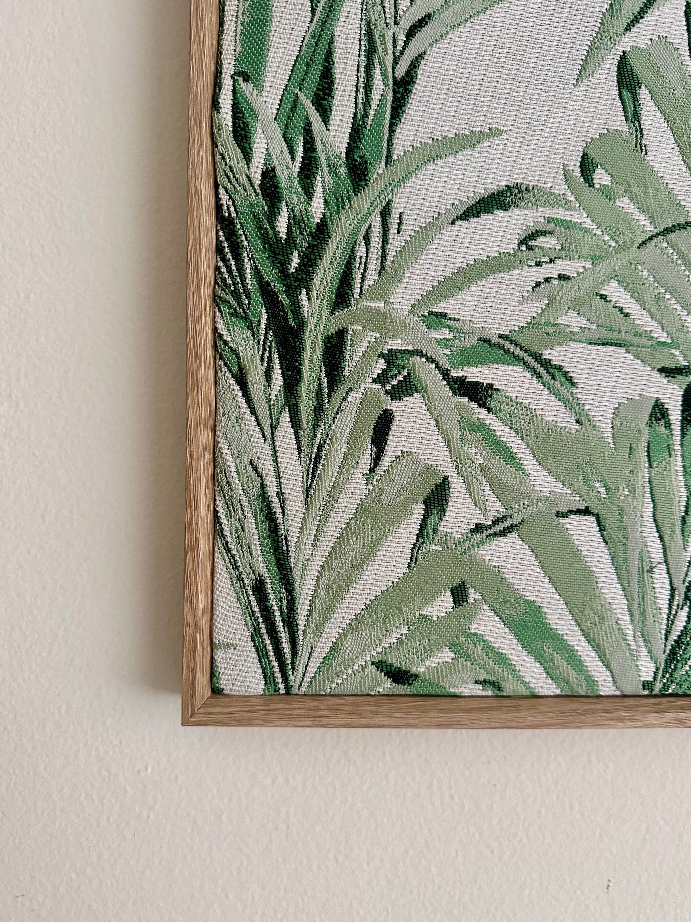 A close-up of framed botanical wall art showing green tropical leaves on a light textured background, mounted on a beige wall.