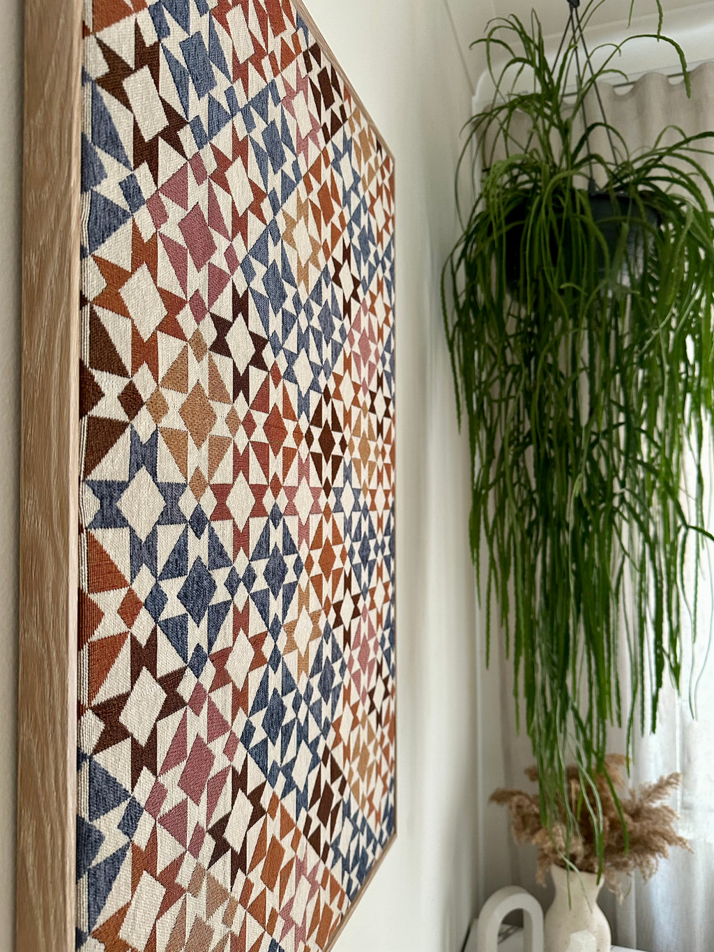 A large square wall art piece with a geometric triangle pattern in earthy tones, hanging on a white wall next to a tall green hanging plant.