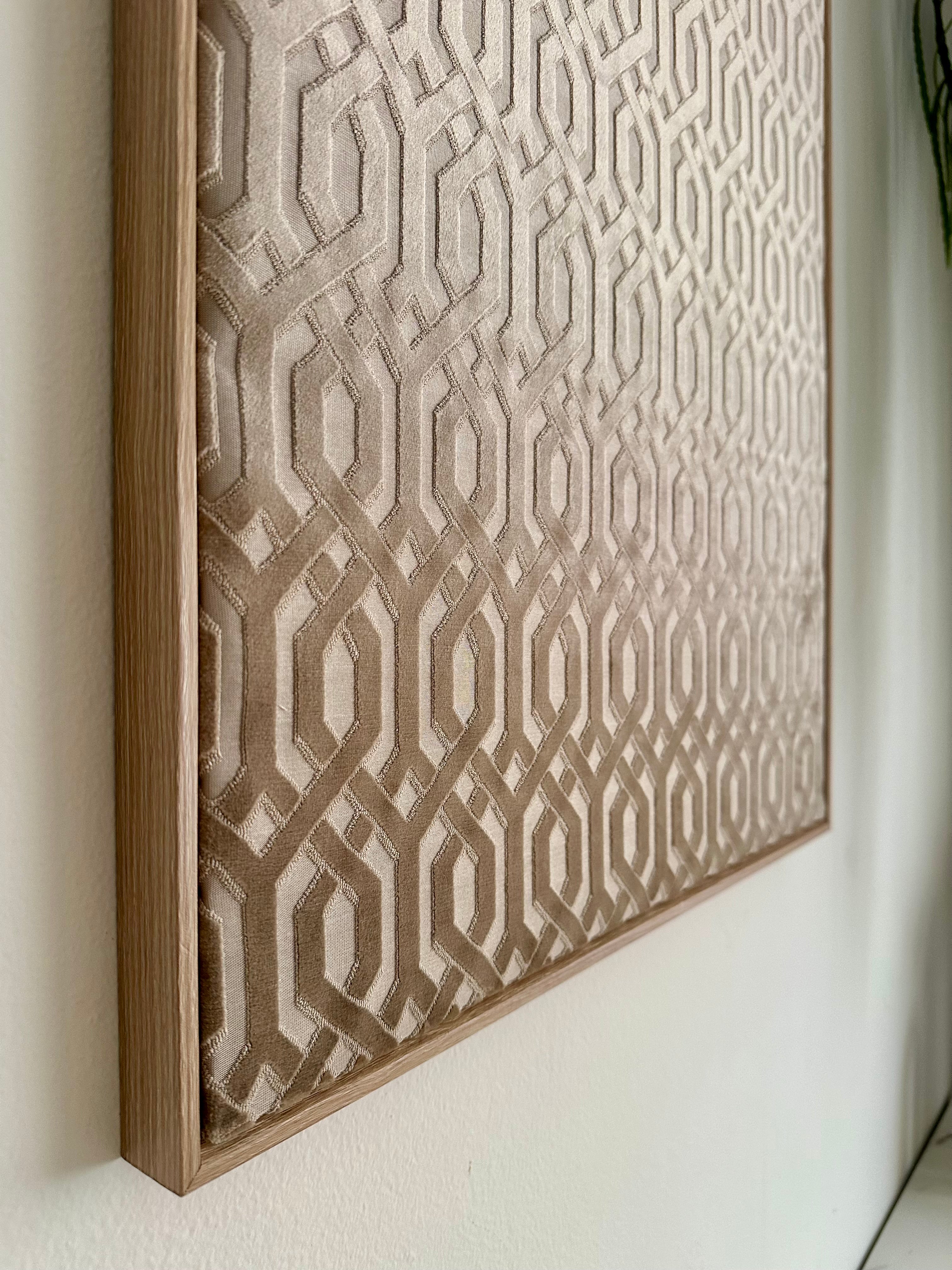 A framed wall art piece with a geometric interlocking pattern in beige and taupe tones, mounted on a white wall.