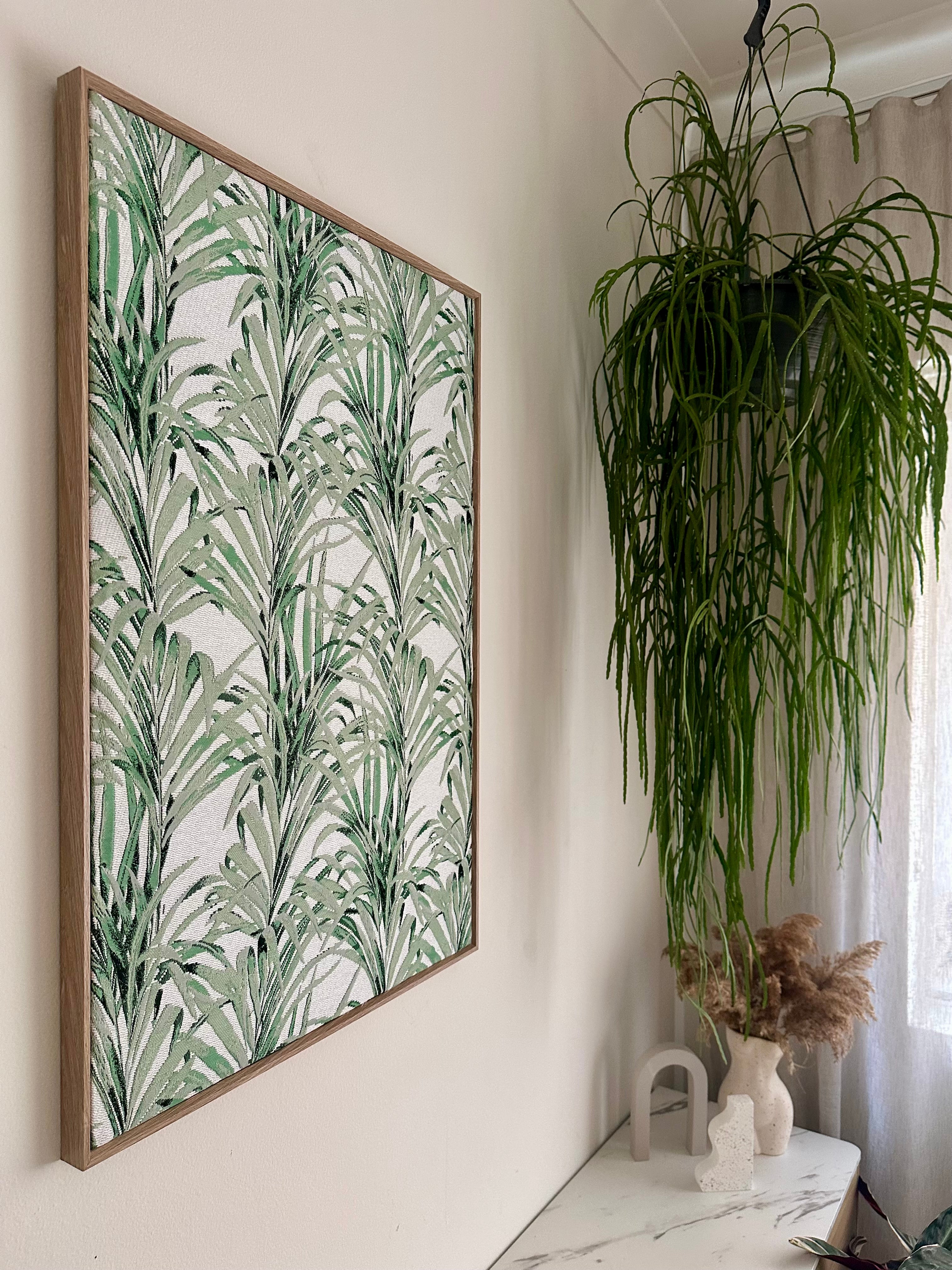 Large framed botanical wall art with a green palm leaf pattern on a white background, hanging on a white wall in a styled interior next to a potted plant and decorative vases.