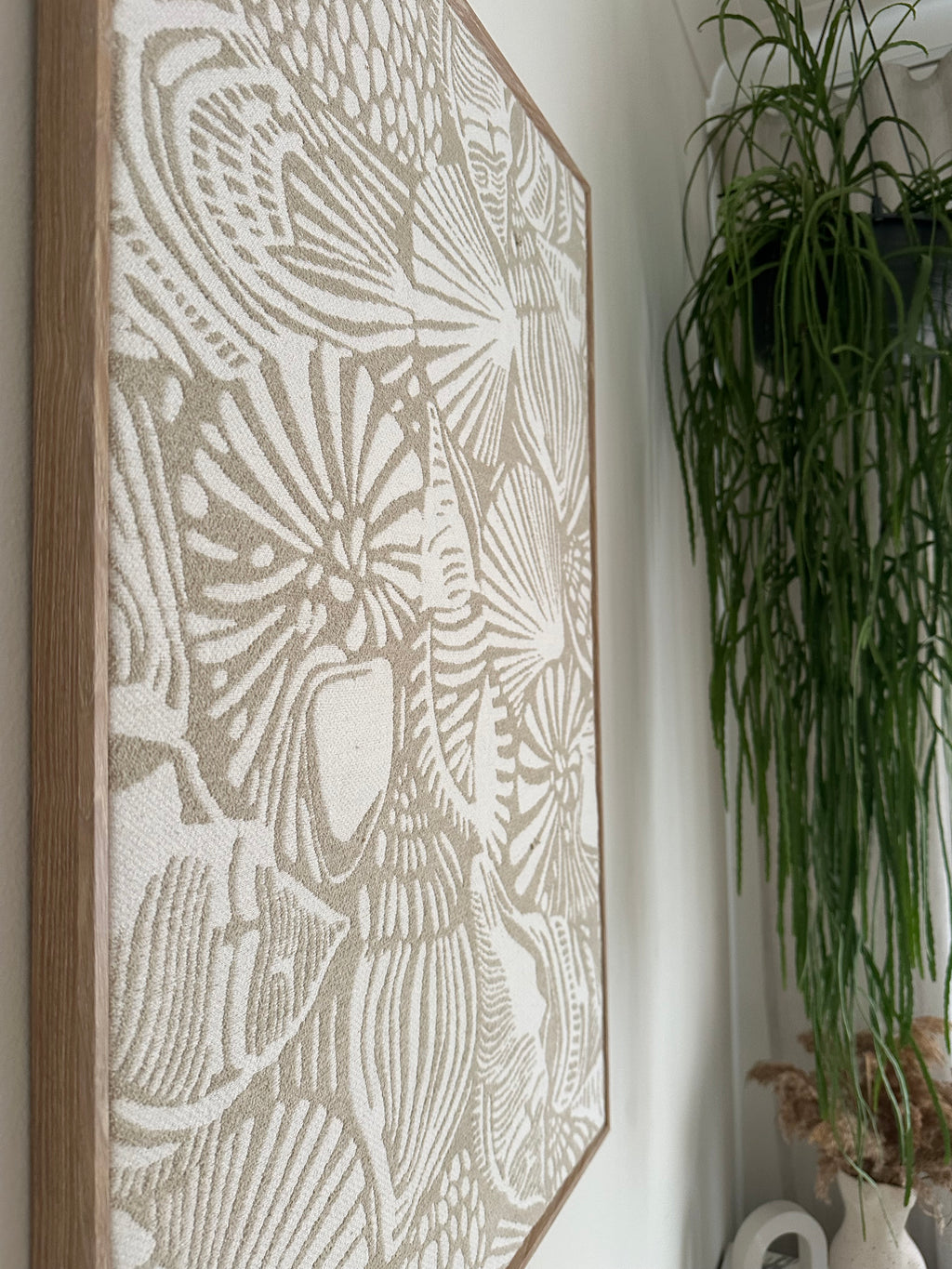 A rectangular framed wall art print featuring a raised, textured botanical leaf pattern in warm beige and off-white, with a natural wood frame, displayed on a white surface with green foliage in the background.