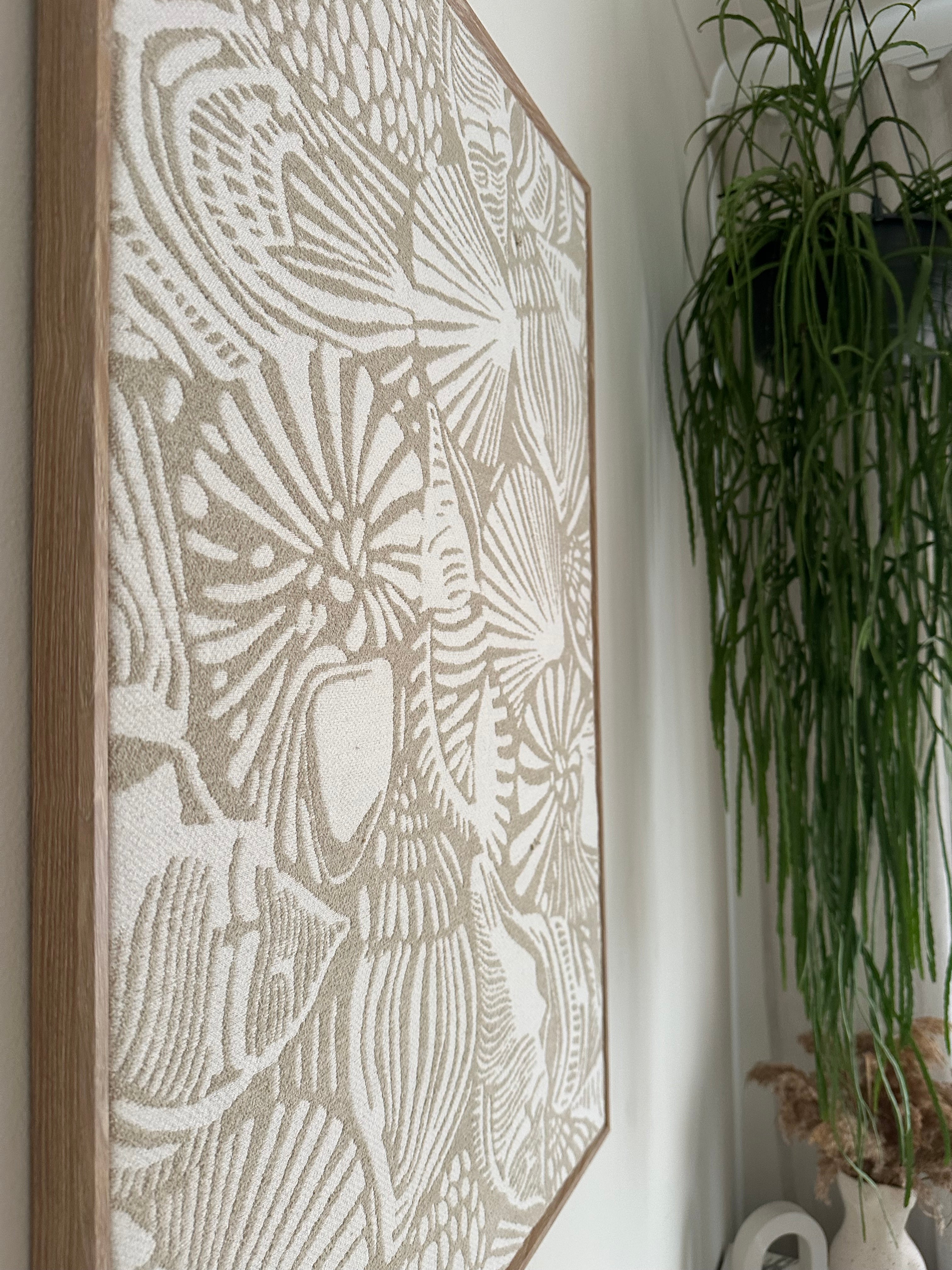 A rectangular framed wall art print featuring a raised, textured botanical leaf pattern in warm beige and off-white, with a natural wood frame, displayed on a white surface with green foliage in the background.