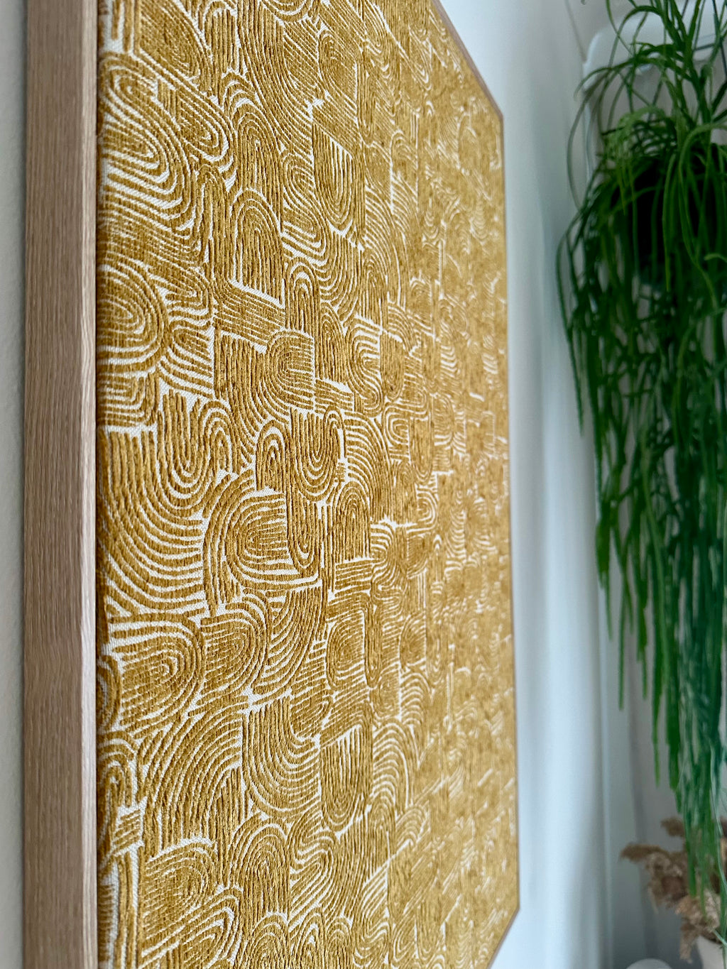 A large framed wall art piece with a textured golden-yellow and white abstract pattern, shown hanging on a white wall next to a green hanging plant.