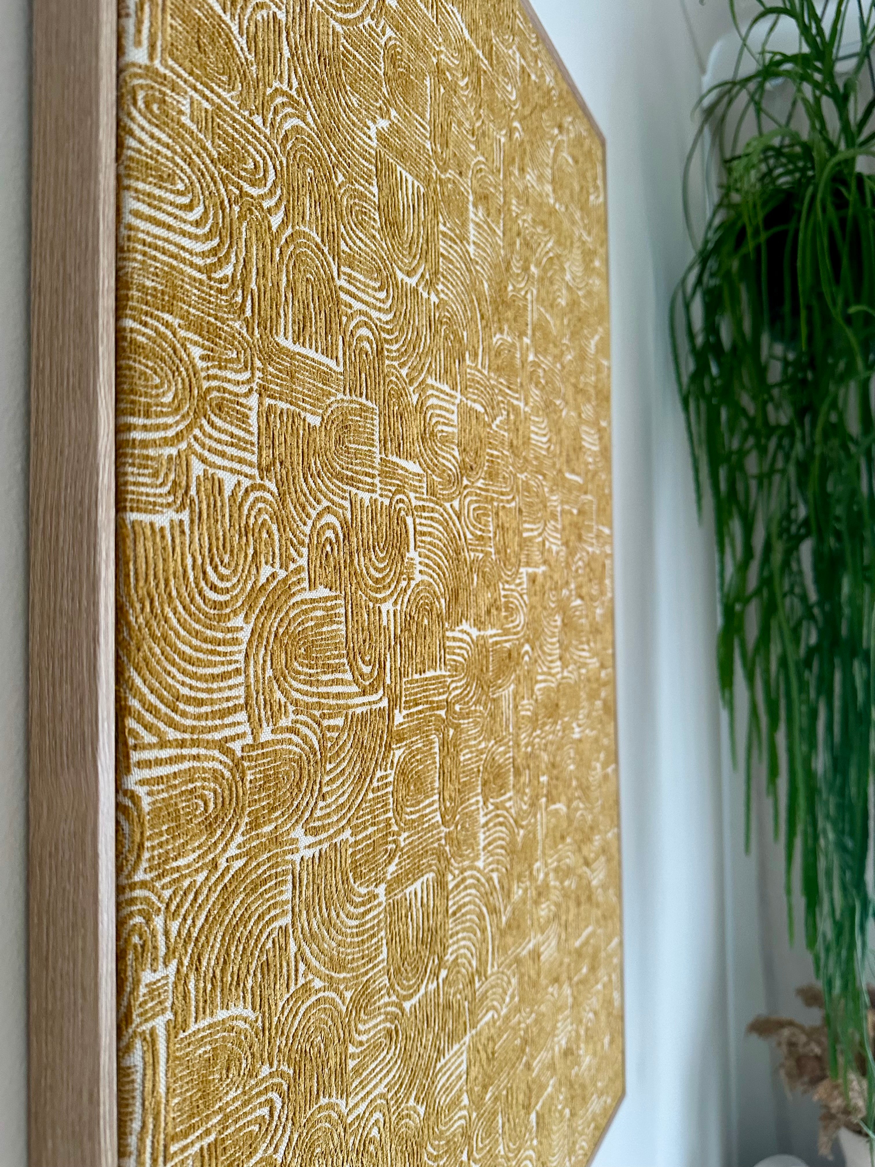 A large framed wall art piece with a textured golden-yellow and white abstract pattern, shown hanging on a white wall next to a green hanging plant.