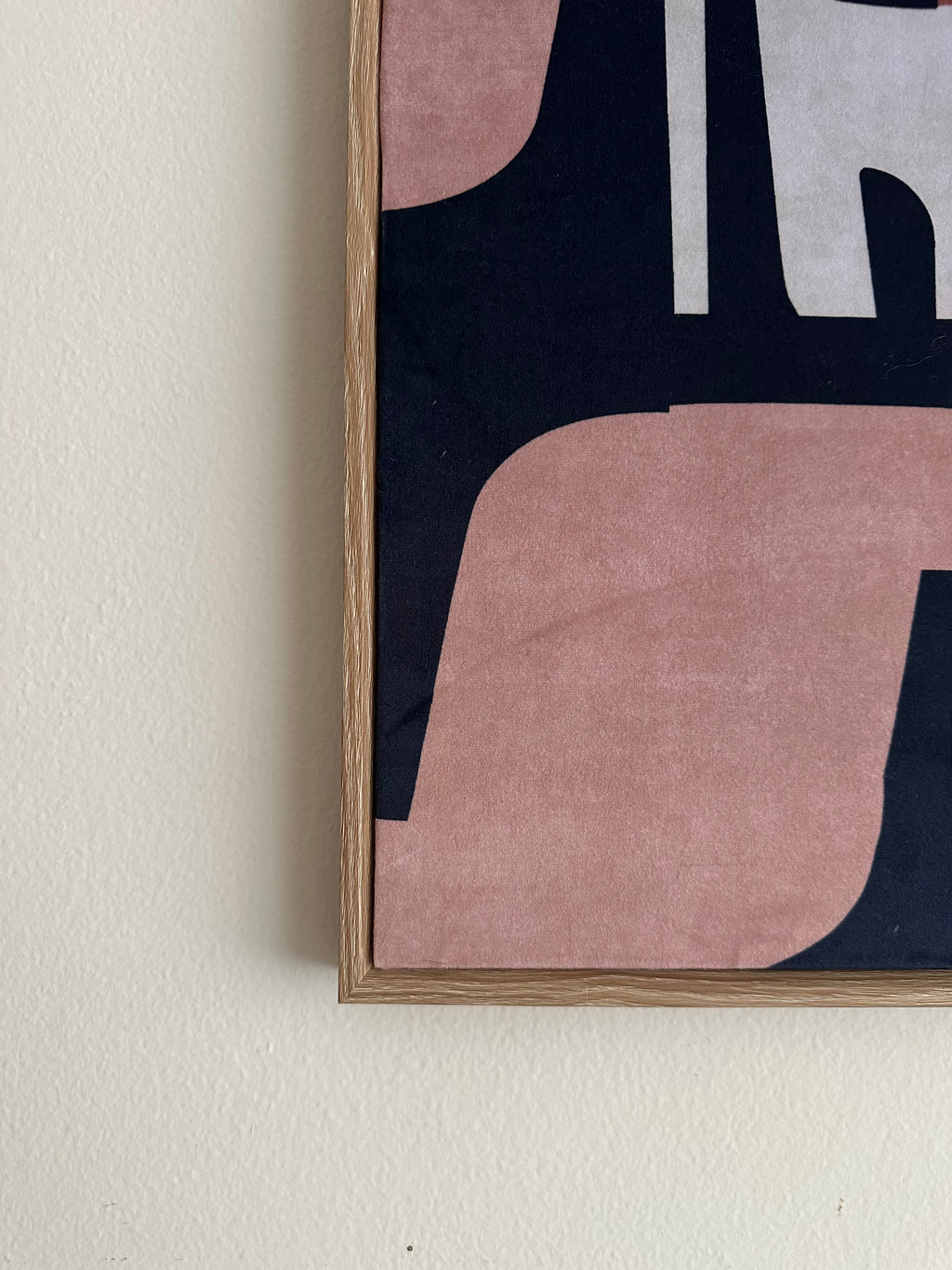 A close-up of framed wall art hanging on a light beige wall, showing an abstract design with dusty pink, navy blue, and white shapes inside a light wood frame.