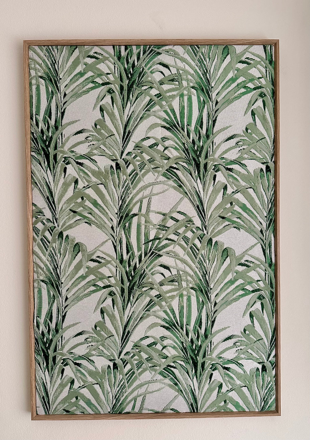 A framed botanical art print featuring a green palm leaf pattern on a textured off-white background, enclosed in a light brown wooden frame and mounted on a plain beige wall.