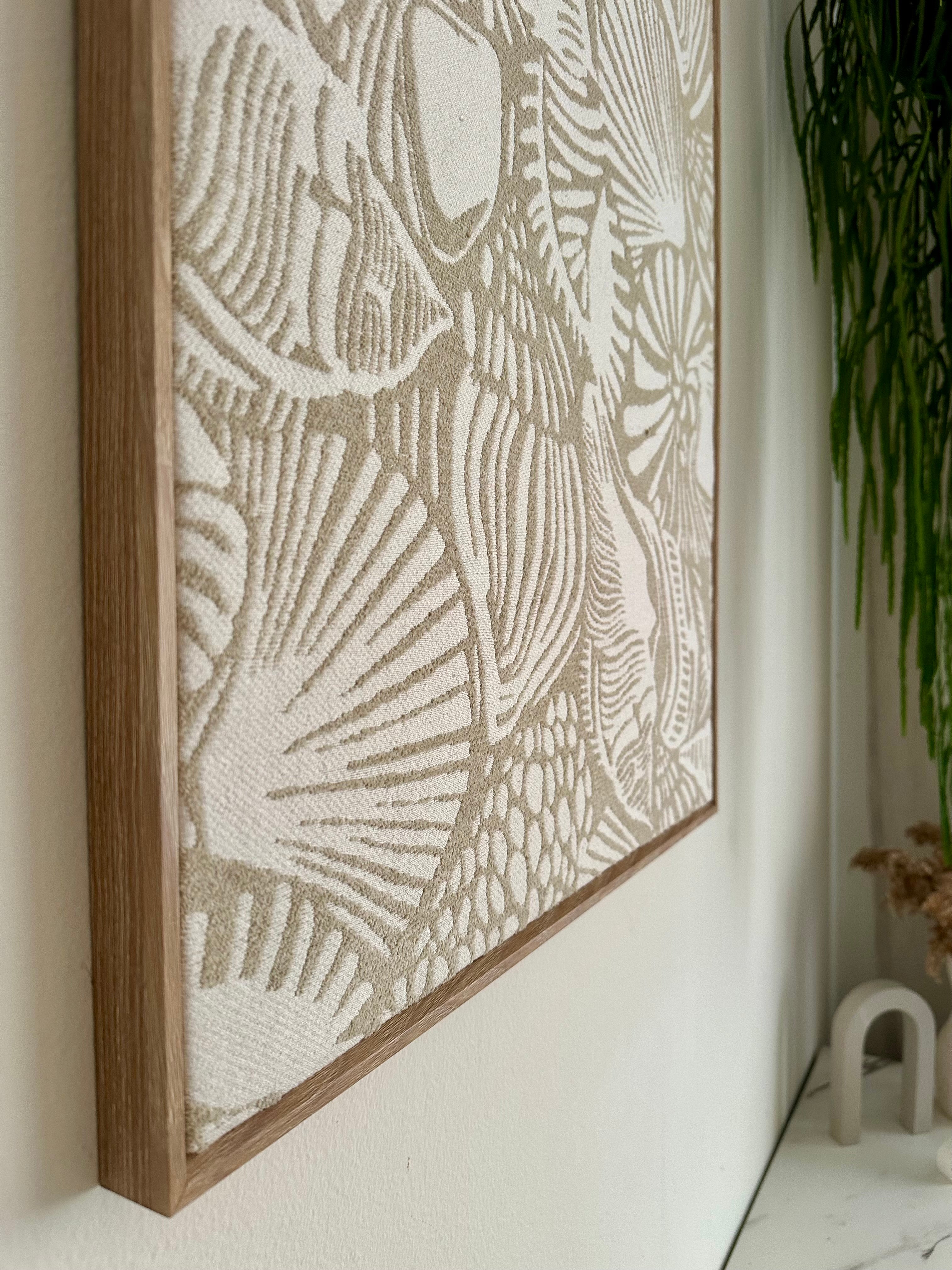 A framed wall art piece with an abstract woven or textured pattern in beige and off-white tones, shown hanging on a white wall in a styled interior with a green plant and small decor visible.