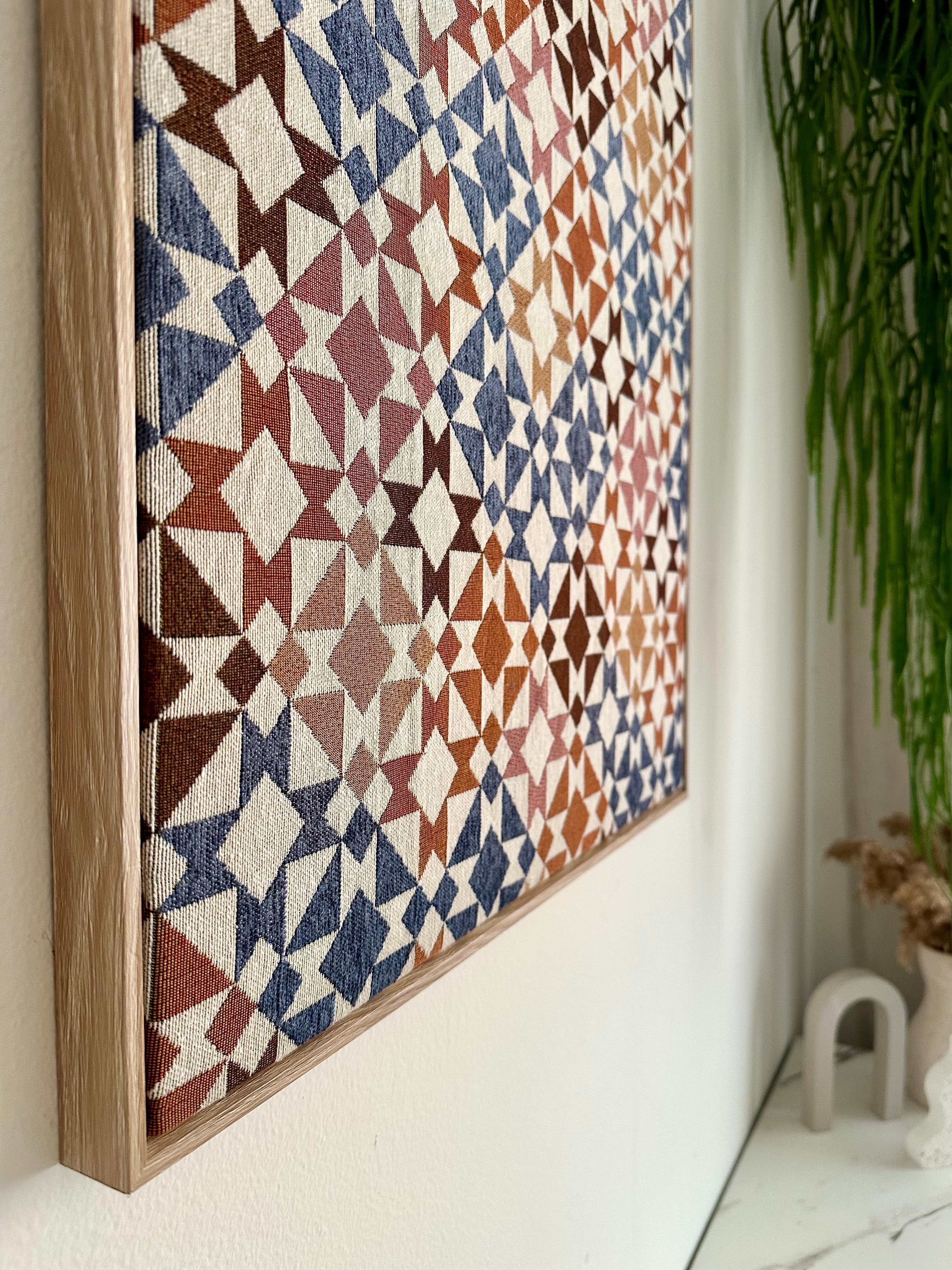 A framed geometric wall art piece with a woven textile pattern in rust, blue, cream, and terracotta, hanging on a white wall next to a green plant.