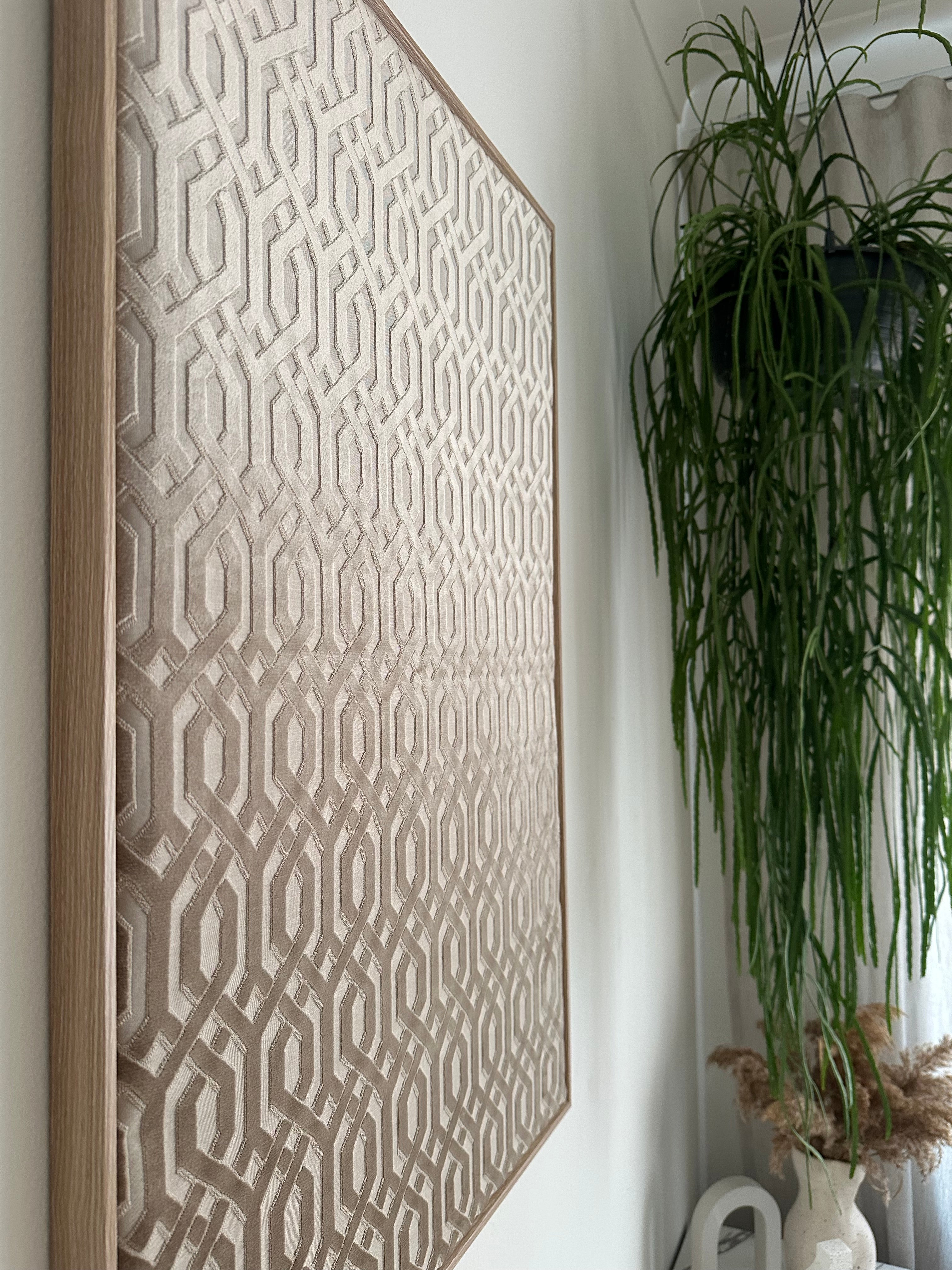A rectangular area rug with a geometric interlocking pattern in beige and taupe tones, featuring a light natural wood frame, displayed on a white surface with a green plant in the background.